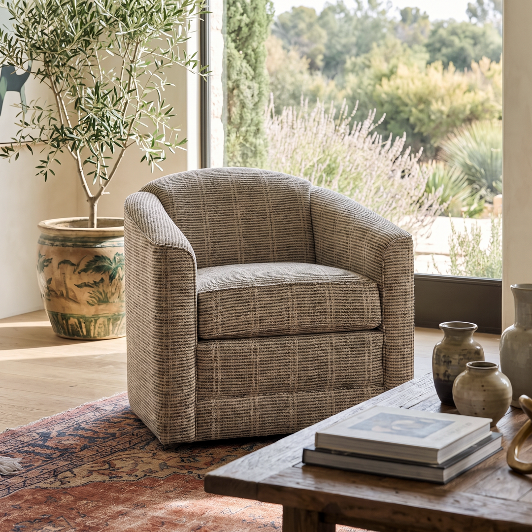 Cozy living room featuring a plaid barrel chair, olive tree, and rustic wooden coffee table in a serene, sunlit space.