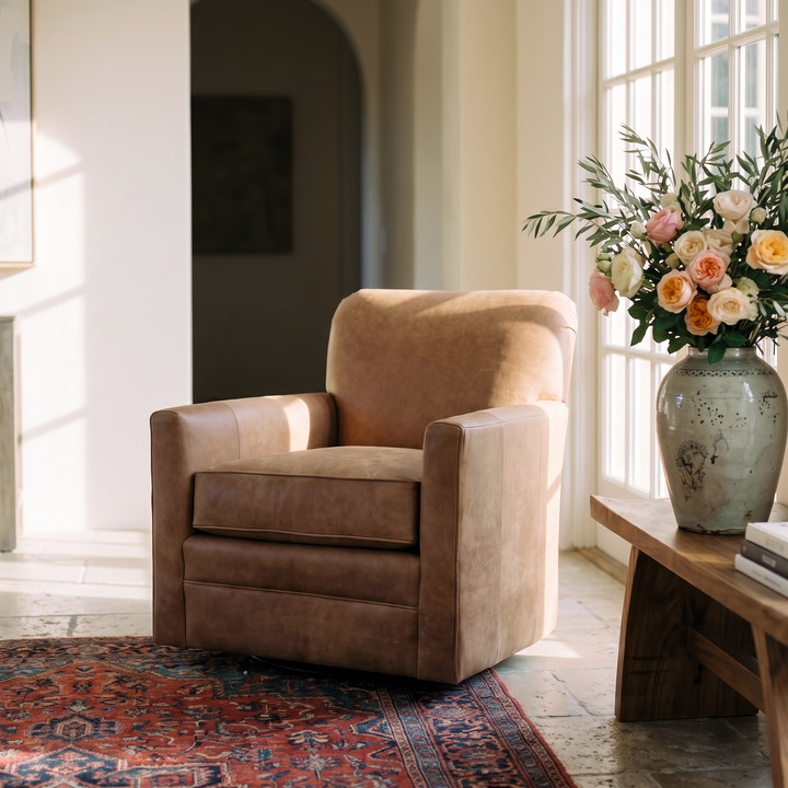 A cozy, sunlit living room featuring a tan leather swivel chair, a vintage-style rug, and a floral vase on a wooden side table.