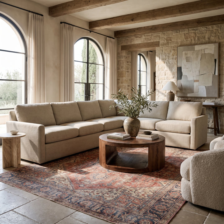 Modern rustic living room with a beige sectional sofa, round wood coffee table, and patterned rug in a sunlit stone space.