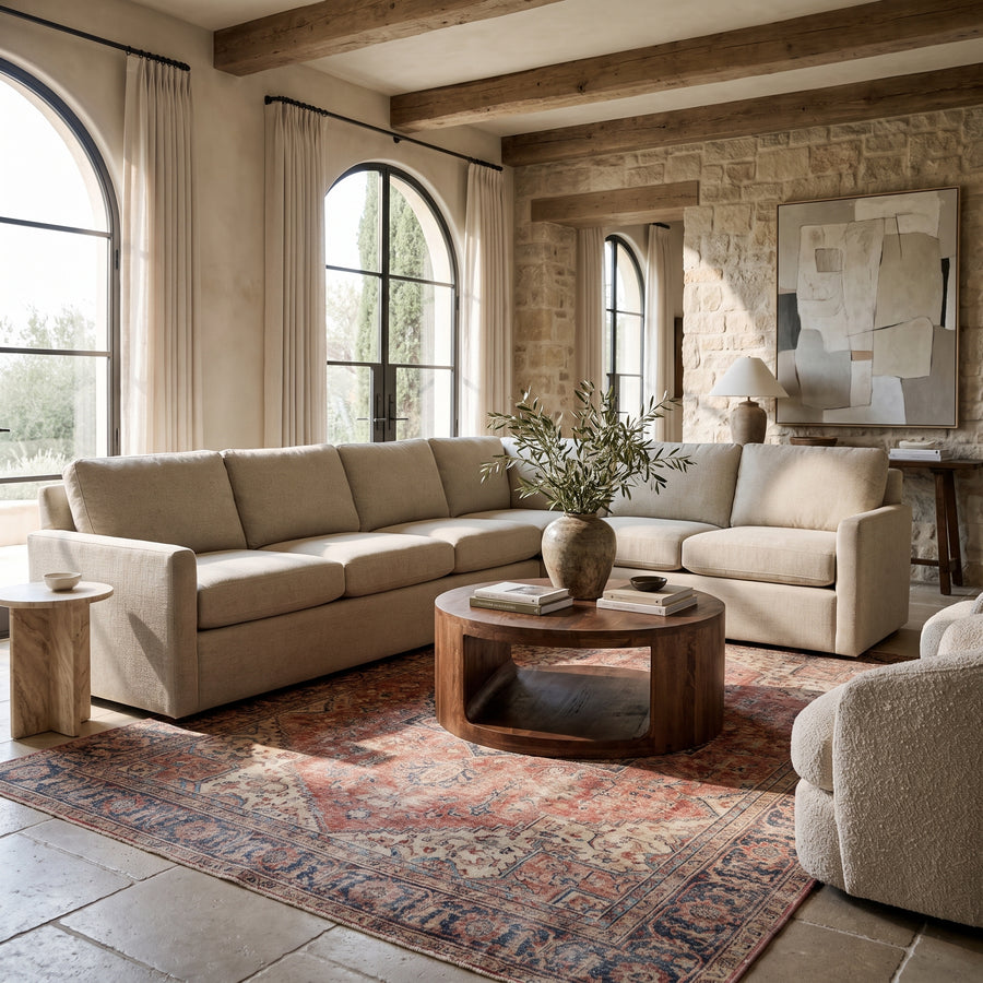 Modern rustic living room with a beige sectional sofa, round wood coffee table, and patterned rug in a sunlit stone space.