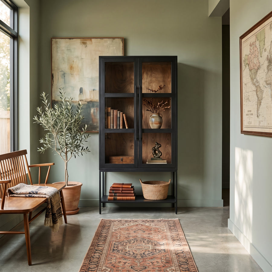 Modern entryway with olive tree, wooden bench, black glass display cabinet, and vintage rug in a sage green room.