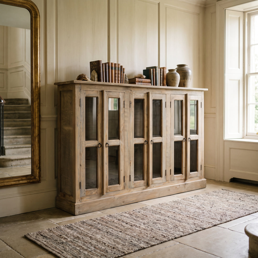 Rustic wooden cabinet with glass doors in a classic, light-filled room with a gold-framed mirror and stone flooring.