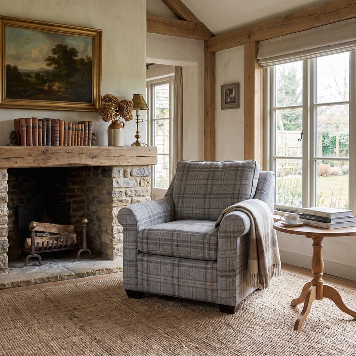 Cozy rustic living room featuring a plaid armchair, stone fireplace, wooden beams, and a bright window with garden views.
