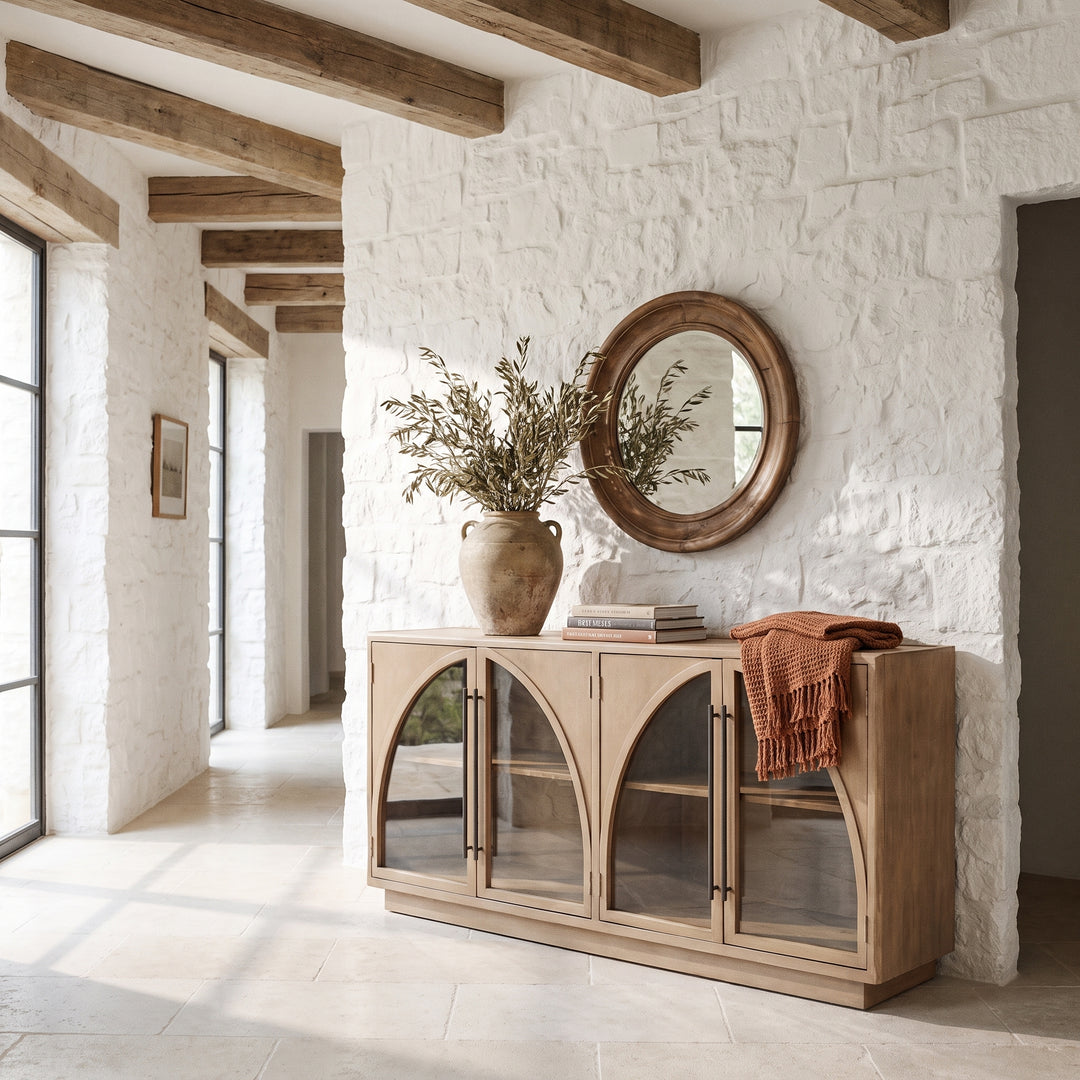 Rustic entryway with white stone walls, a wooden arched cabinet, round mirror, and olive branches in a ceramic vase.